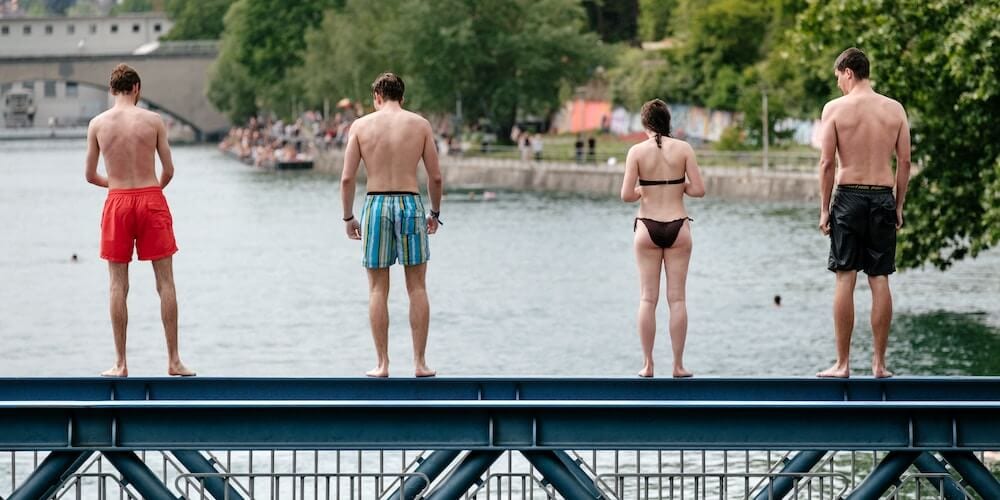 Vier Personen in Badebekleidung stehen mit dem Rücken zum Wasser auf einem Steg.